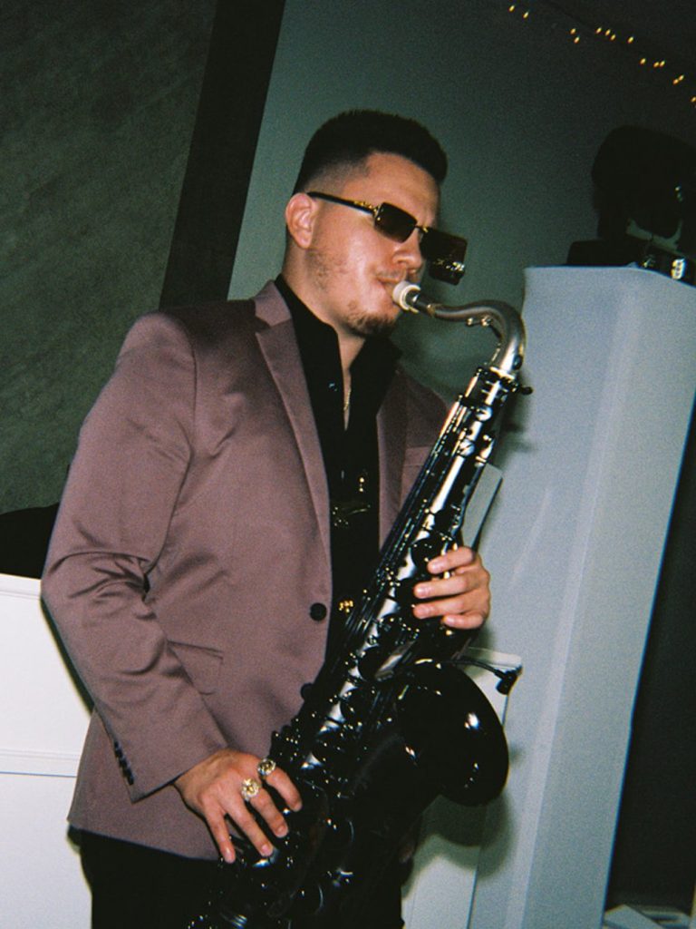 Male musician wearing a suit and sunglasses playing a black saxophone during the wedding reception.