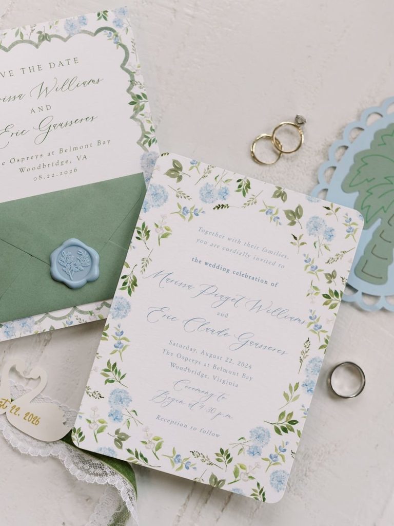Flat lay of a wedding invitation suite featuring a white card with a blue floral border, a sage green envelope with a light blue wax seal, and three wedding rings resting nearby.