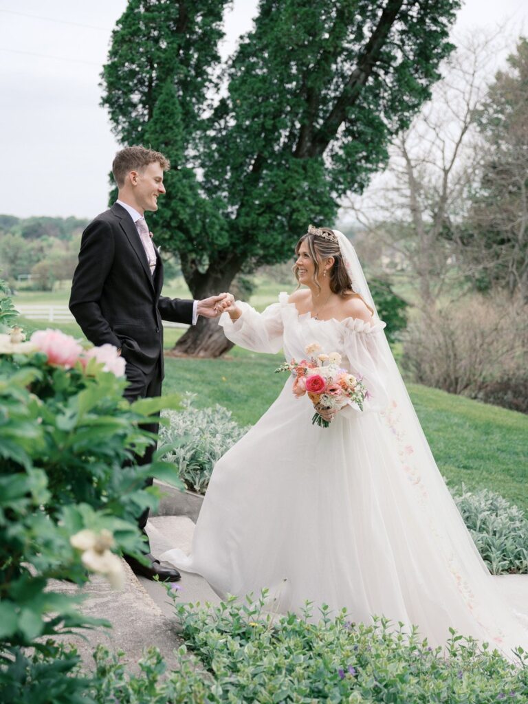 Groom Colin holding hands with bride Tori as she walks past him outdoors in a white off-the-shoulder wedding gown, holding a bouquet of pink and peach flowers.