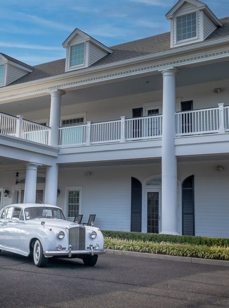Vintage white getaway car parked outside The Carriage House in Galloway, New Jersey, a white colonial-style South Jersey wedding venue with tall columns, a two-story veranda, and dormer windows, ideal for classic car wedding photos.