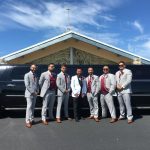 Smiling bride in a lace gown and veil sits with four bridesmaids in shimmering champagne-gold gowns inside a wedding party bus. They all stretch out their hands to show off their rings and manicures, a fun getaway moment for South Jersey weddings and bridal party transportation.
