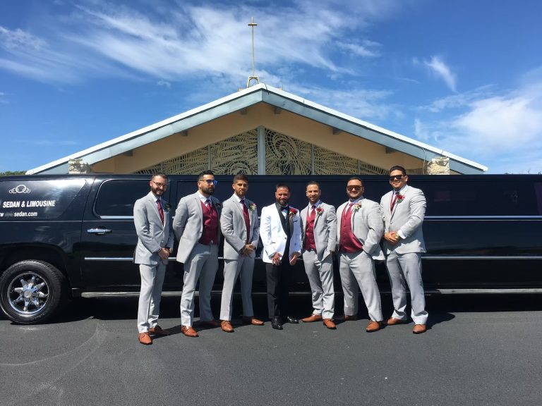 Smiling bride in a lace gown and veil sits with four bridesmaids in shimmering champagne-gold gowns inside a wedding party bus. They all stretch out their hands to show off their rings and manicures, a fun getaway moment for South Jersey weddings and bridal party transportation.