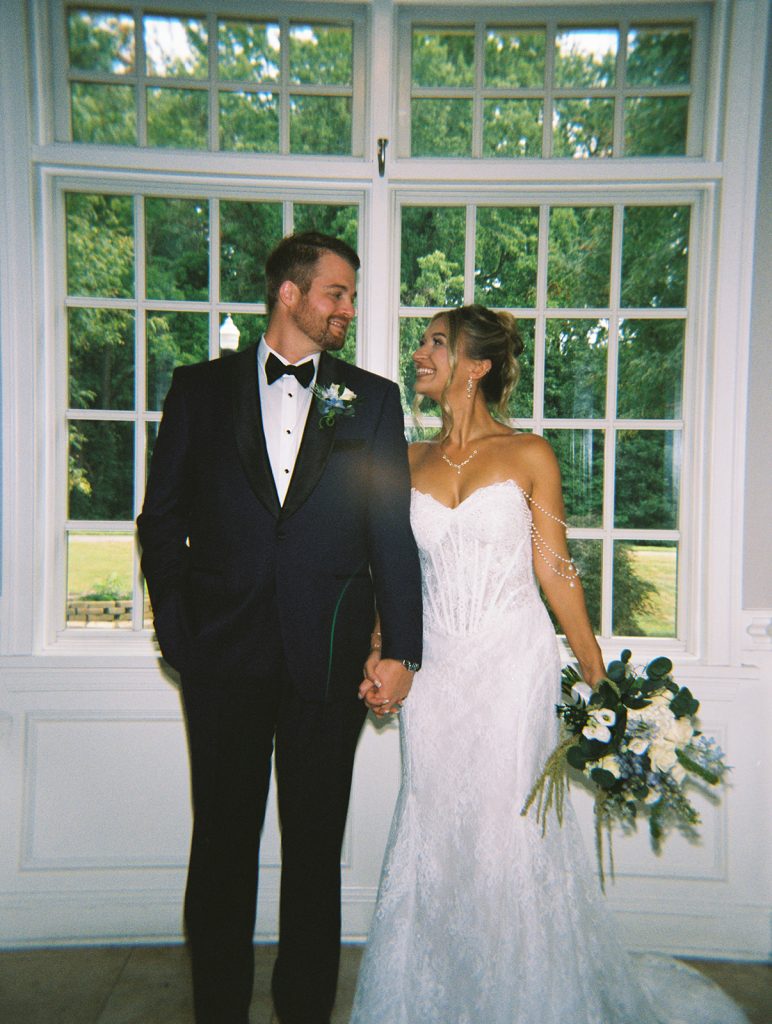 Bride and groom holding hands and smiling at each other in front of a grid-pane window indoors.