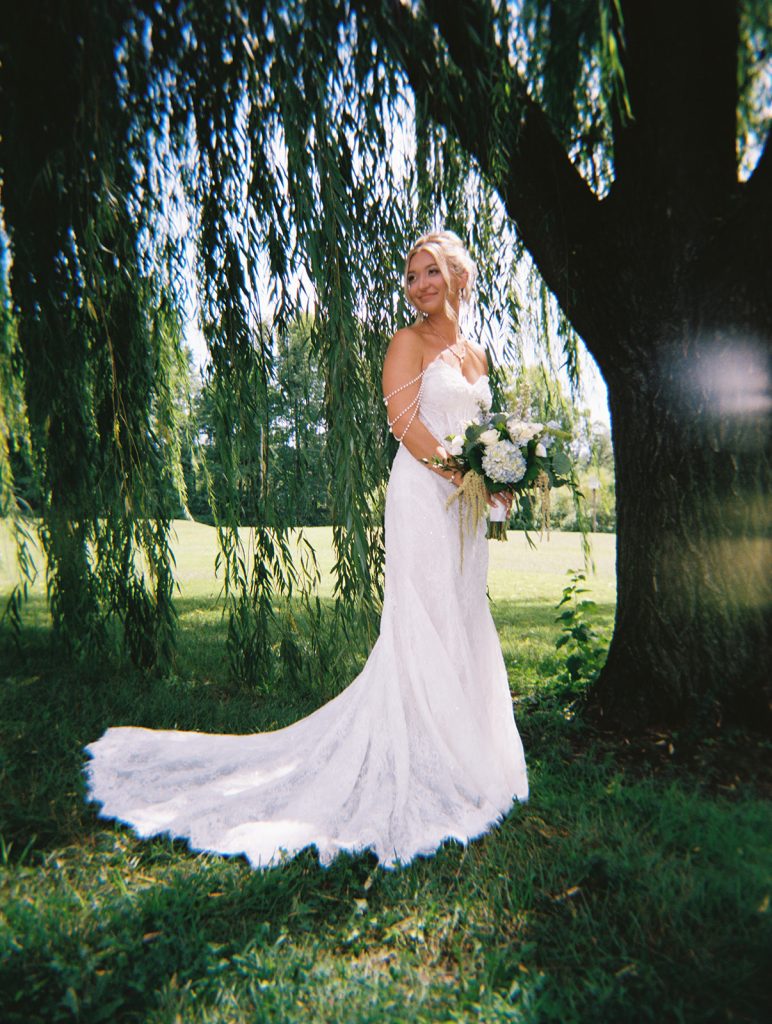 Bride in a white gown holding a bouquet while standing beneath the weeping branches of a large tree outdoors.