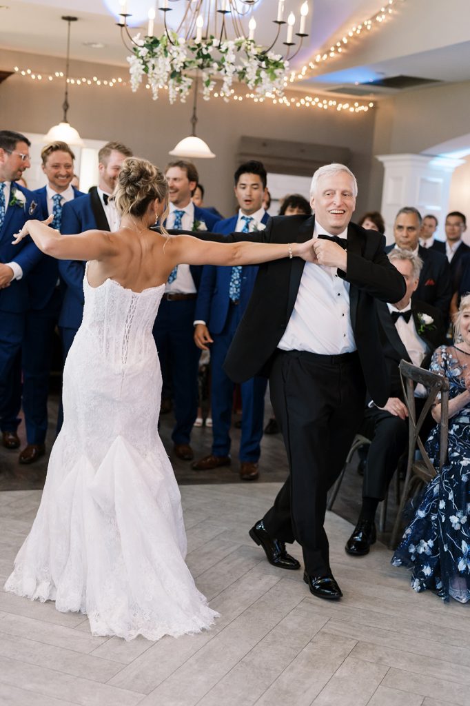 Bride in a white wedding dress dancing with a man in a black tuxedo during the reception, while guests look on and smile.